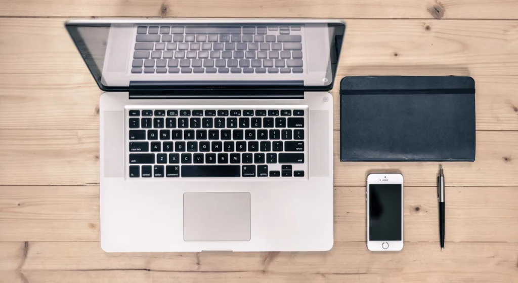 Top view of a tidy workspace featuring a laptop, smartphone, notebook, and pen on a wooden desk