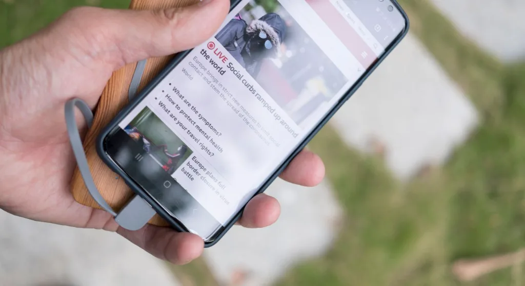 A hand holds a smartphone displaying online news while charging with a portable power bank